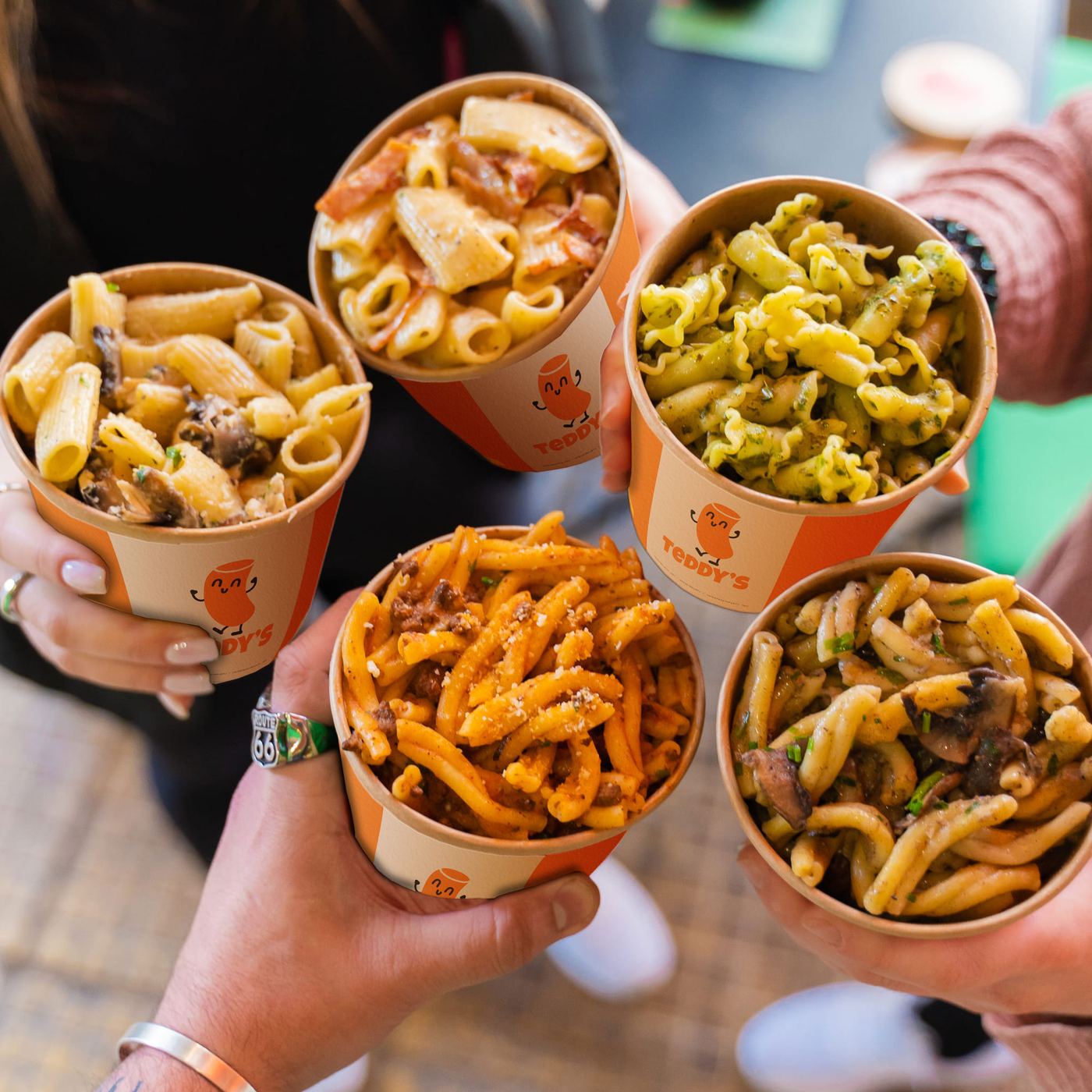Five hands holding Teddy's pasta cups together — carbonara, pesto, bolognese, truffle mushroom and tomato
