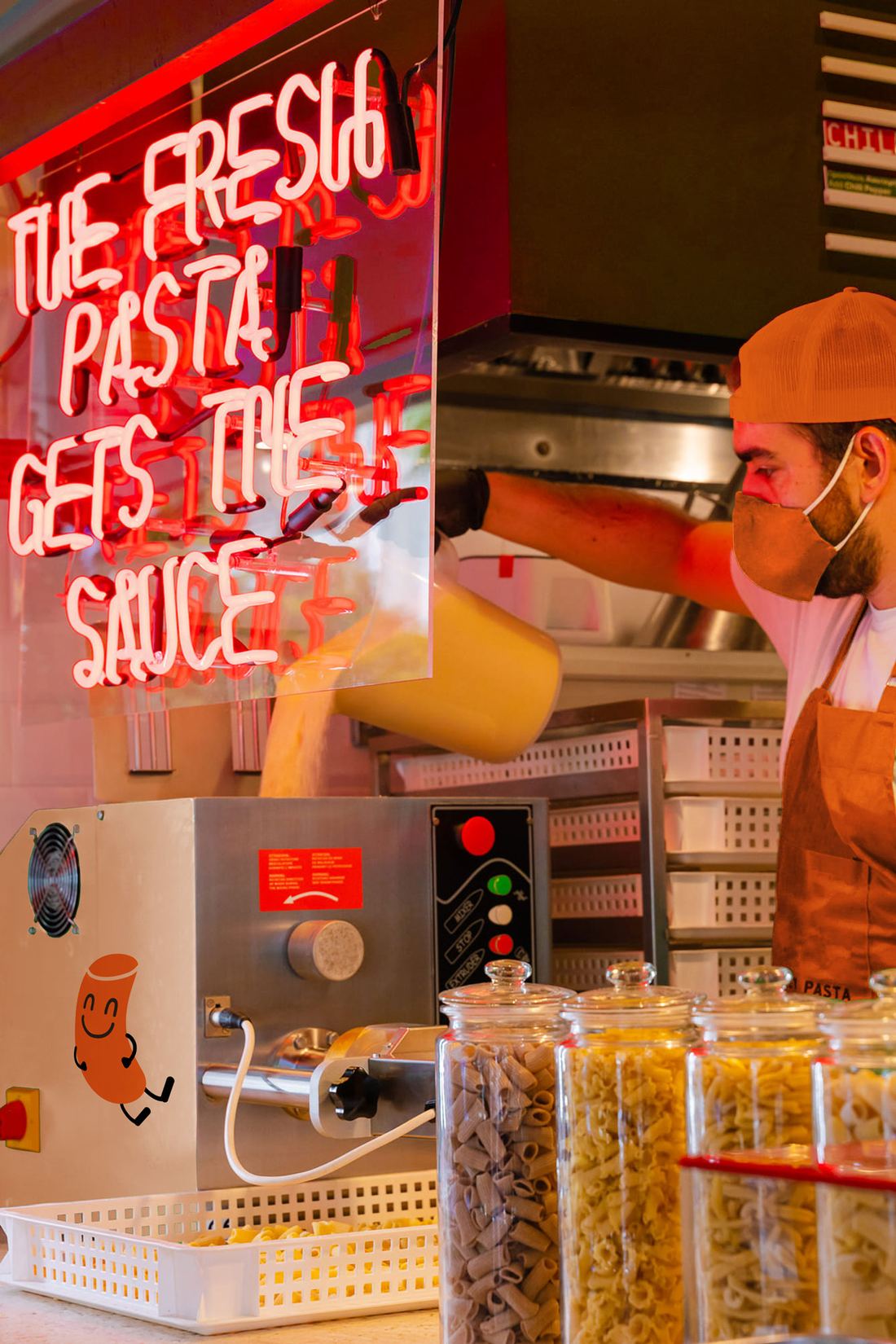 Fresh pasta coming out of the extruder beneath a neon sign reading 'The fresh pasta gets the sauce'